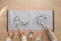 Two children paint side by side on the long grey Water Canvas with plain water, wooden rods at each end: a house and stick family on one side, toddler scribbles on the other