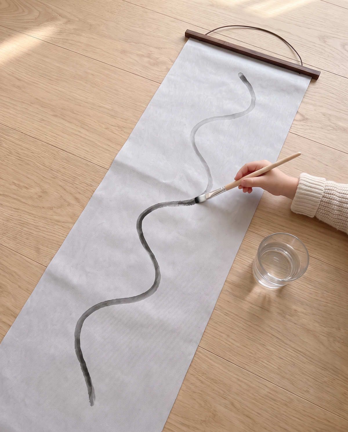 A small hand paints one long wavy line with plain water down the grey Water Canvas on an oak floor, a dark wooden rod and cord at the top, a glass of water beside it